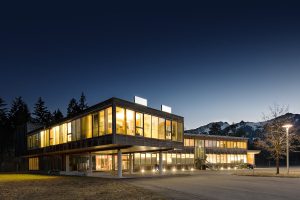 At dusk, a contemporary, well-lit two-storey MySalon Suite showcases expansive glass windows set against a mountainous backdrop, with trees surrounding the scene.