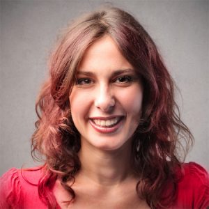With long, wavy auburn hair and a bright smile, a person stands confidently in a red top against a grey backdrop, reminiscent of Kwikshift Removals’ seamless service—effortlessly smooth and expertly managed.