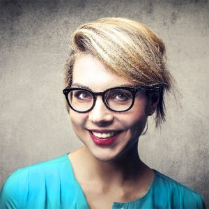 A person with short blonde hair, donning black-framed glasses and a turquoise top, smiles at the camera against a textured grey background. It feels like a snapshot captured just before they set off on a new adventure with Kwikshift Removals.